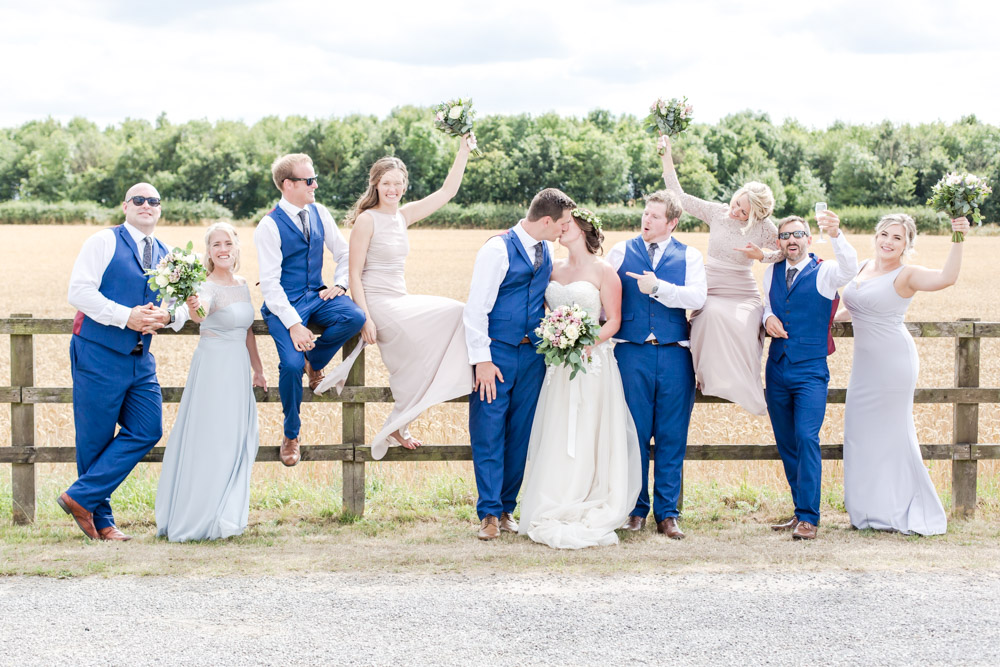 grooms men and bridesmaids sat on a fence cheering for the bride and groom kissing