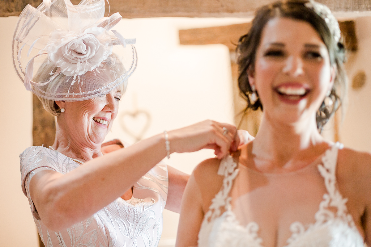 mother of bride fastening wedding dress bride is laughing