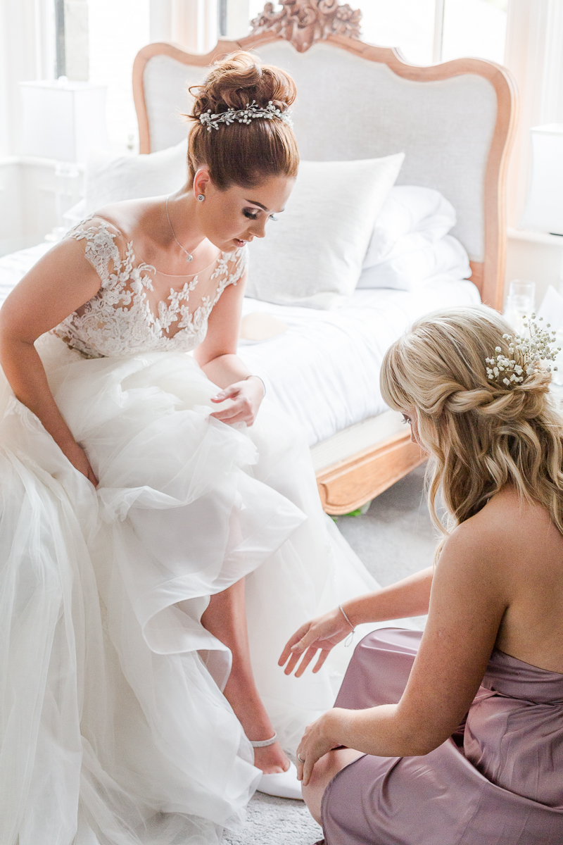 bride sat on edge of bed while bridesmaid helps put shoes on