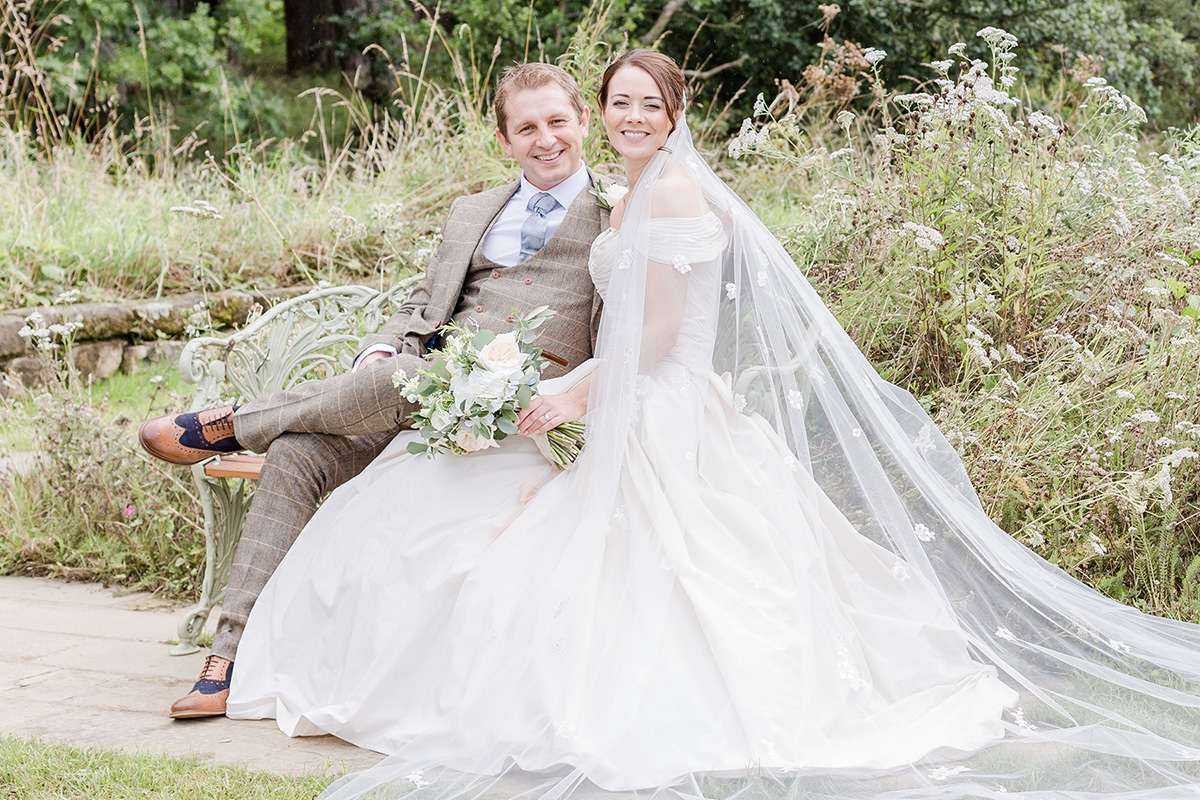 full length gown spilled out onto grass from bench with bride and groom