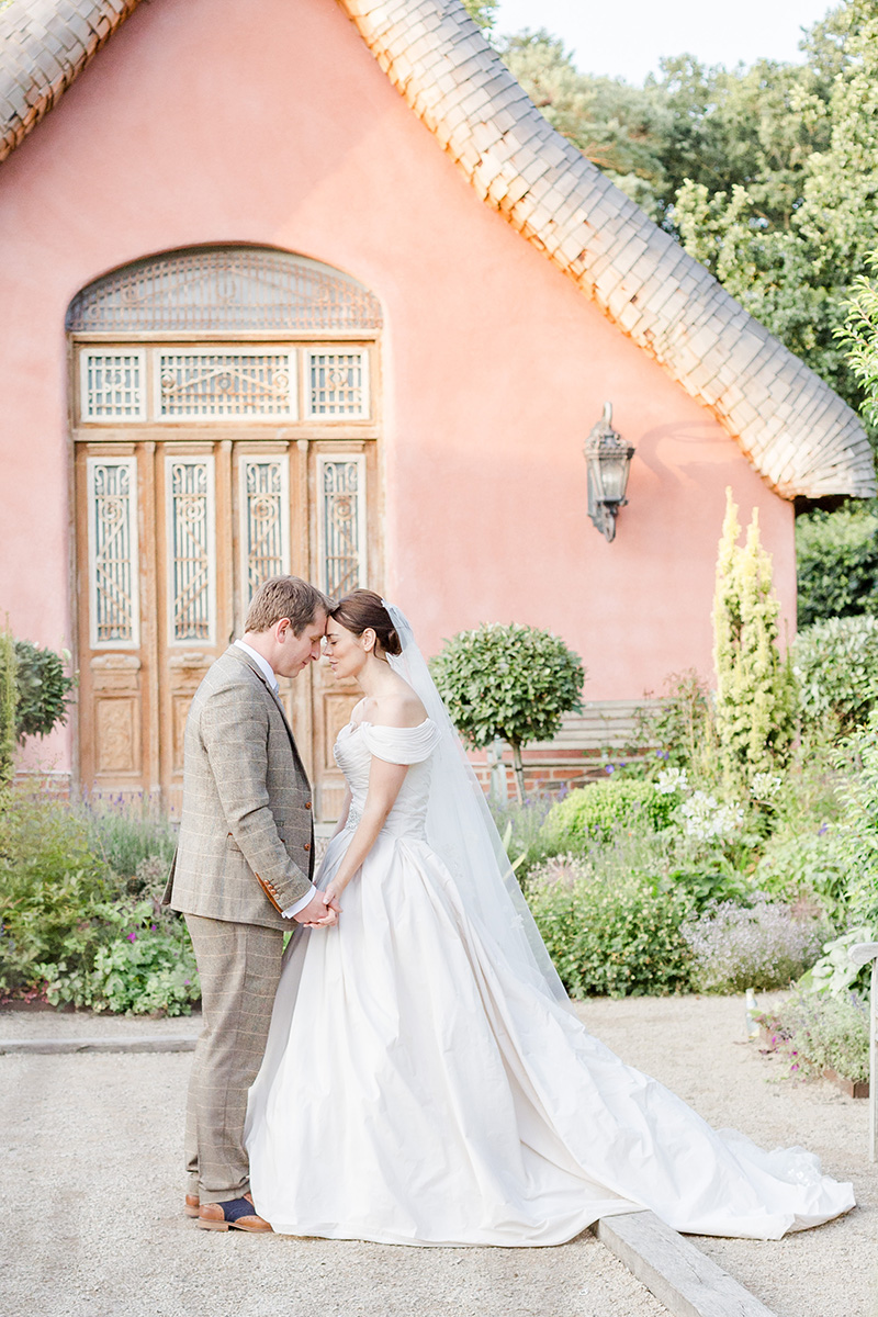 bide and groom forehead to forehead in gardens with pink house behind