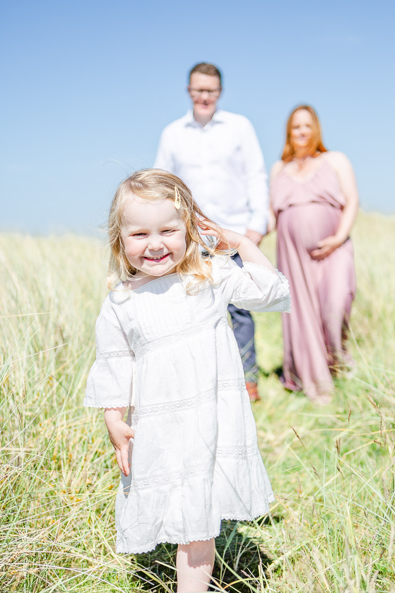 toddler smiling at camera with parents in back