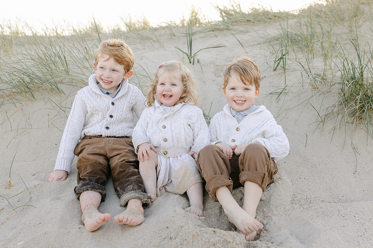 three siblings sat on dunes covered in sand smiling for family photo shoot