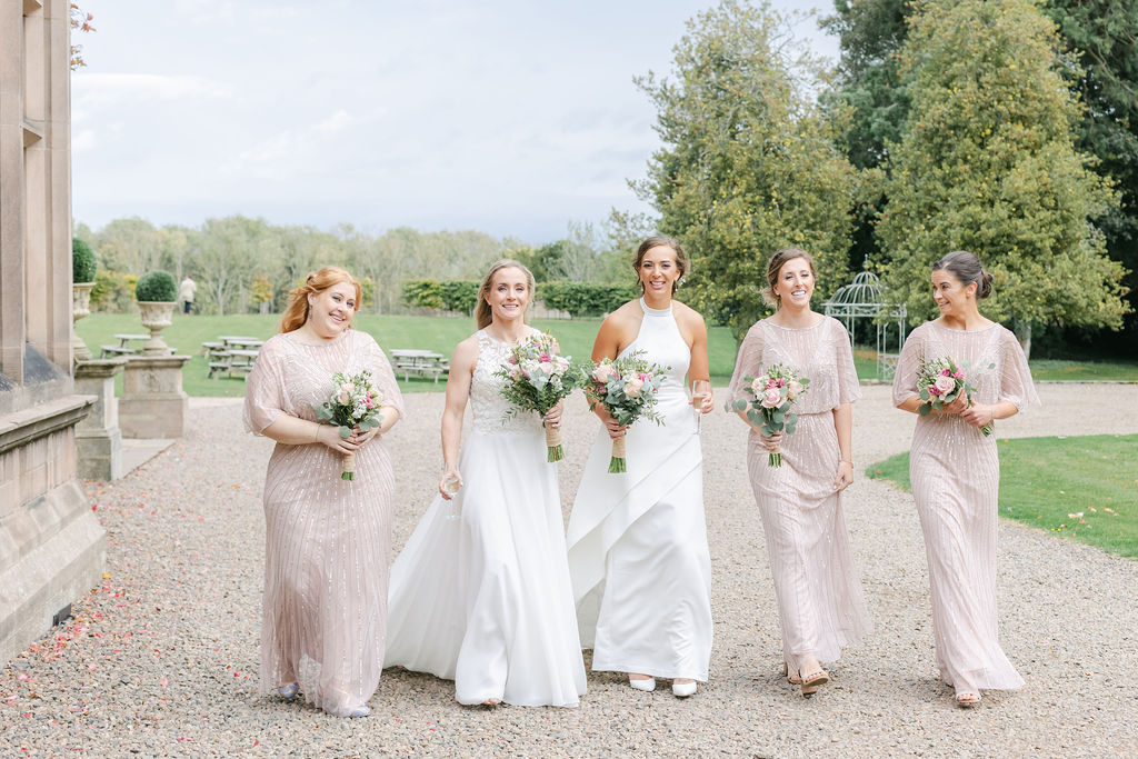 two brides and 3 bridesmaids walking through wedding venue grounds